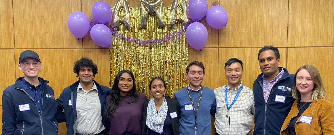 Medical School Students pose in front of a wall with purple and gold balloons that spell out "AMA"