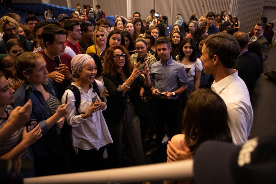 A group of people within a lecture hall surround Beto O’Rourke as he is photographed 
