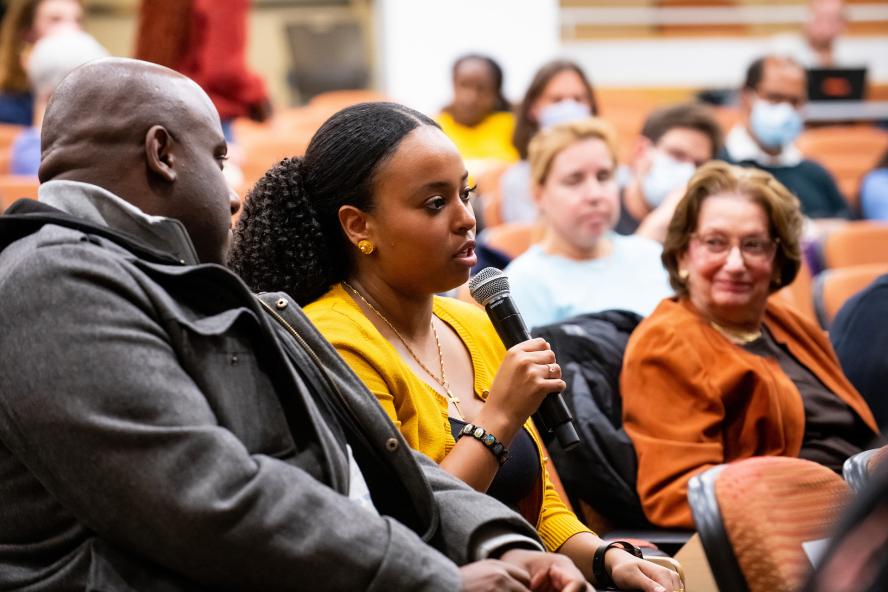 A woman from the audience, wearing a yellow cardigan, is speaking into the microphone