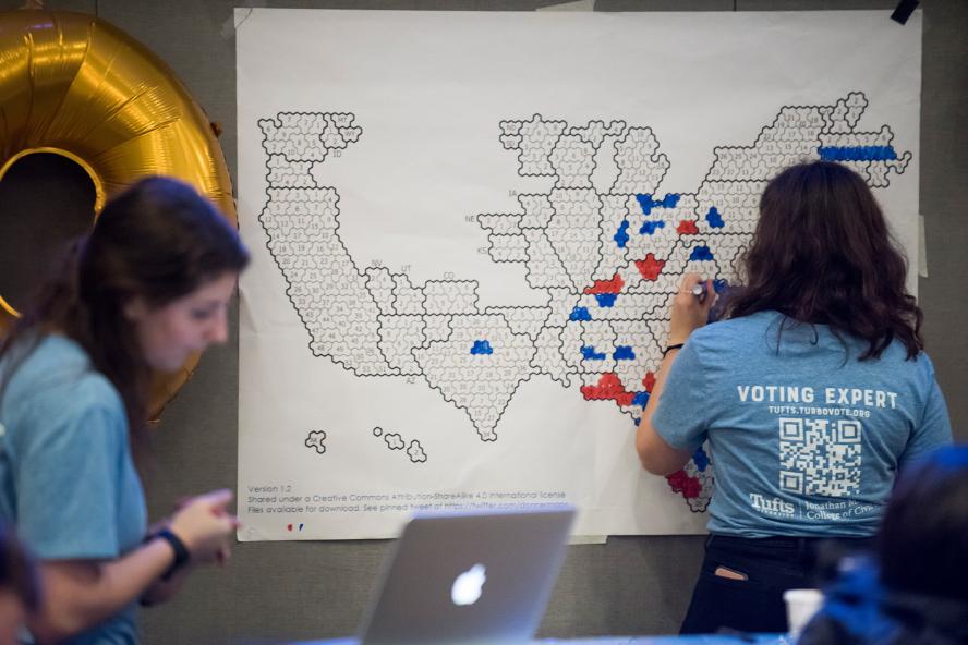 A woman wearing a t-shirt that reads "voting expert" is coloring in areas of a map-like diagram in red and blue