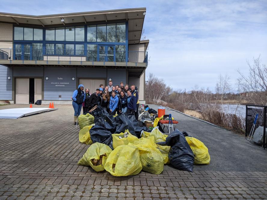 Tufts students pose in front of the Boathouse along the river with garbage bags