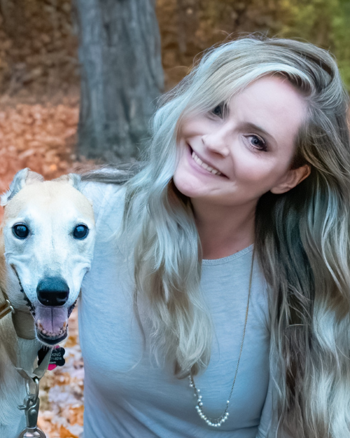 Image of woman smiling with head tilted next to her dog, a greyhound. 