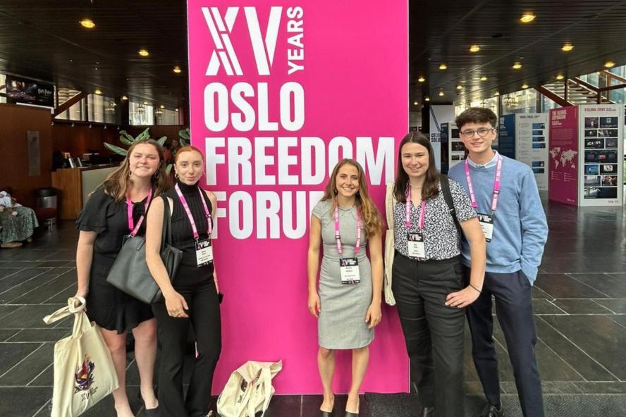 Students stand in front of hot pink Oslo Freedom Forum Sign that reads XV Years