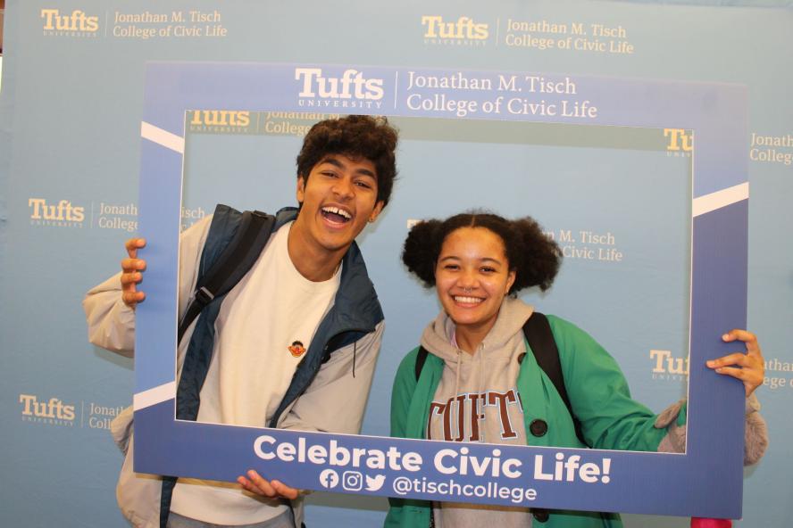 Two students pose with Celebrate Civic Life photo frame in front of blue backdrop with Tisch logo