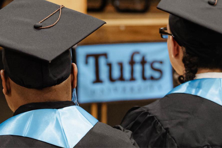 Photo of the back of two graduates in caps and gowns with a blue Tufts University banner in between. 