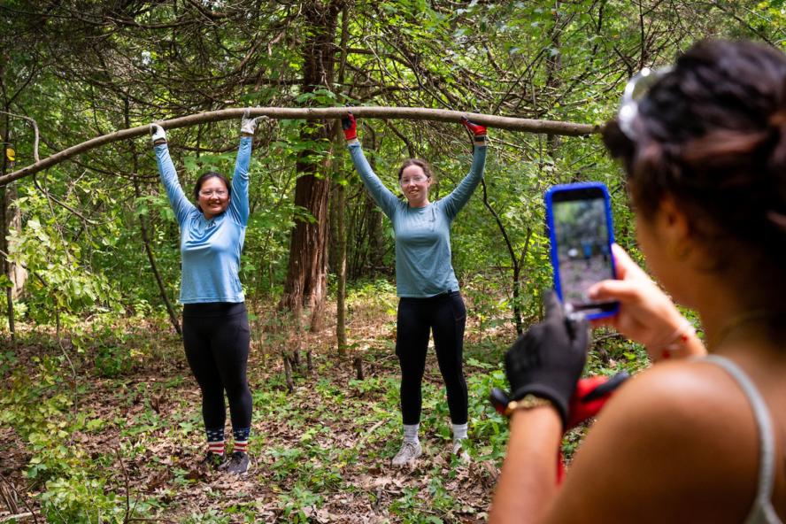 Students hold up a tree branch while a student in the foreground takes a photo on an iPhone