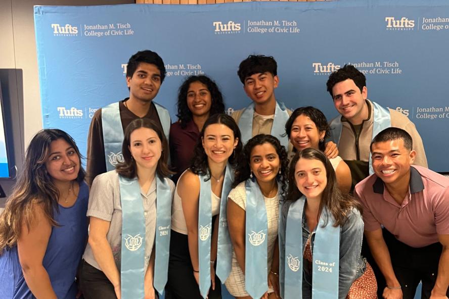 Students wearing graduation cords pose in front of a background with the Tisch College logo