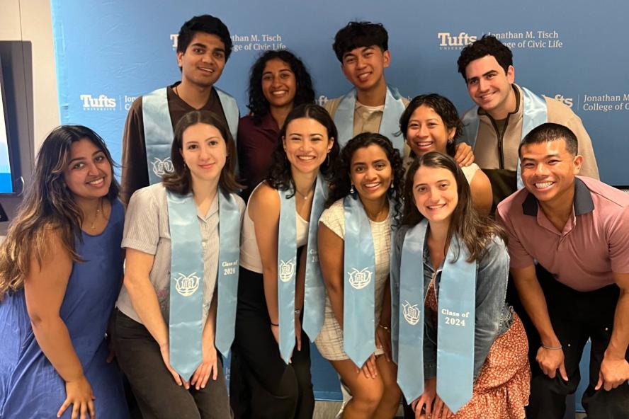 Students wearing graduation cords pose in front of a background with the Tisch College logo