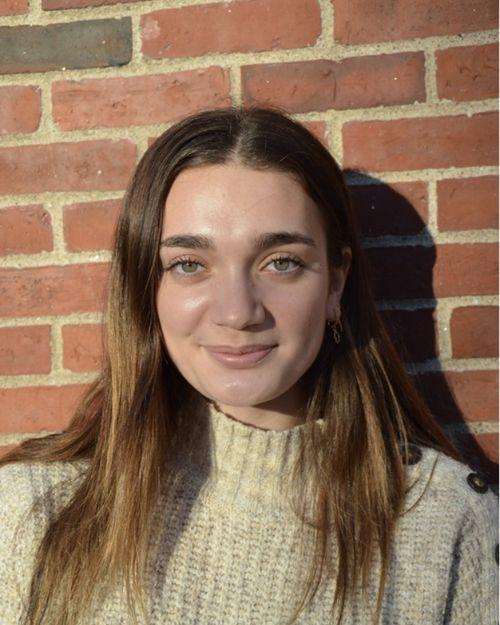 Young woman against a brick background looks straight ahead and smiles.