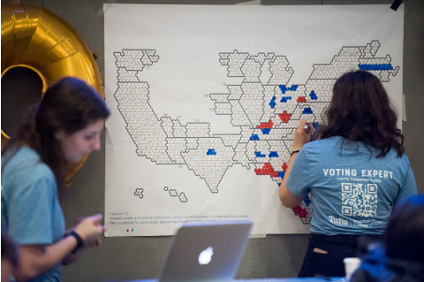 A woman wearing a t-shirt that reads "voting expert" is coloring in areas of a map-like diagram in red and blue