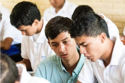 A college student in a blue collared shirt assists a younger student, in a classroom full of students in white uniform shirts.