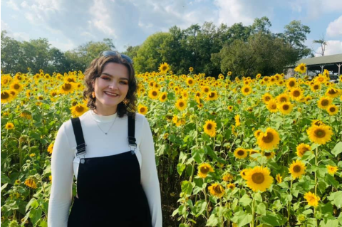 Bethany Kirby in black overalls and a white shirt stands in a field of blooming sunflowers