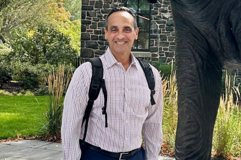 Man in button down and backpack smiles with greenery in background