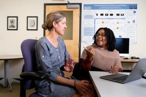 A woman seated at a desk talking with a younger woman, with a laptop open in the foreground. The VERSE summer program teams students new to research with Tufts faculty mentors