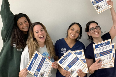Four women in scrubs holding fliers for Tufts Tube videos