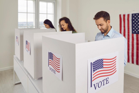 Three young people in voting booths with a U.S. flag in the background. 